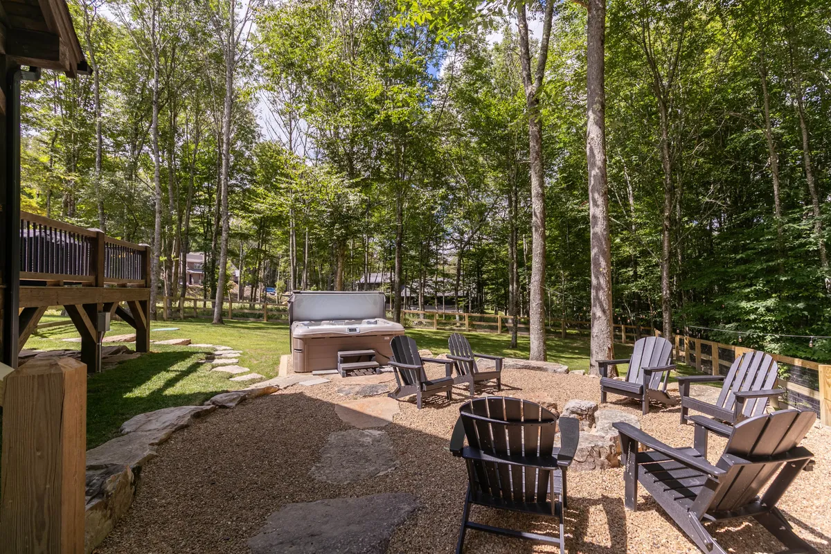 A spacious wooden deck nestled among tall trees, featuring a bubbling hot tub, a fire pit surrounded by adirondack chairs, and inviting spaces for gathering. The fence visible in the background ensures privacy and safety, making it ideal for both relaxing soaks and socializing by the fire.