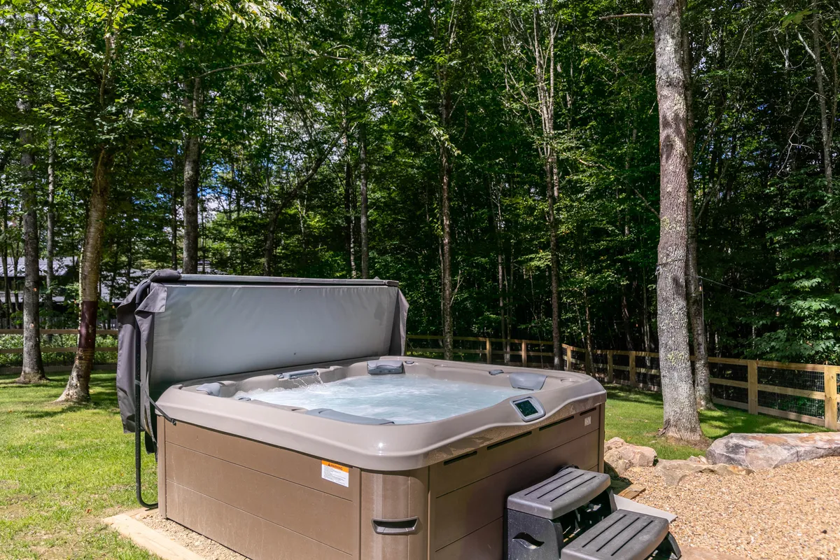 An inviting hot tub sits on a gravel pathway surrounded by trees, offering tranquil views into the wooded landscape. The focus is on outdoor relaxation in nature, with the hot tub as a highlight for unwinding after a day of adventure.