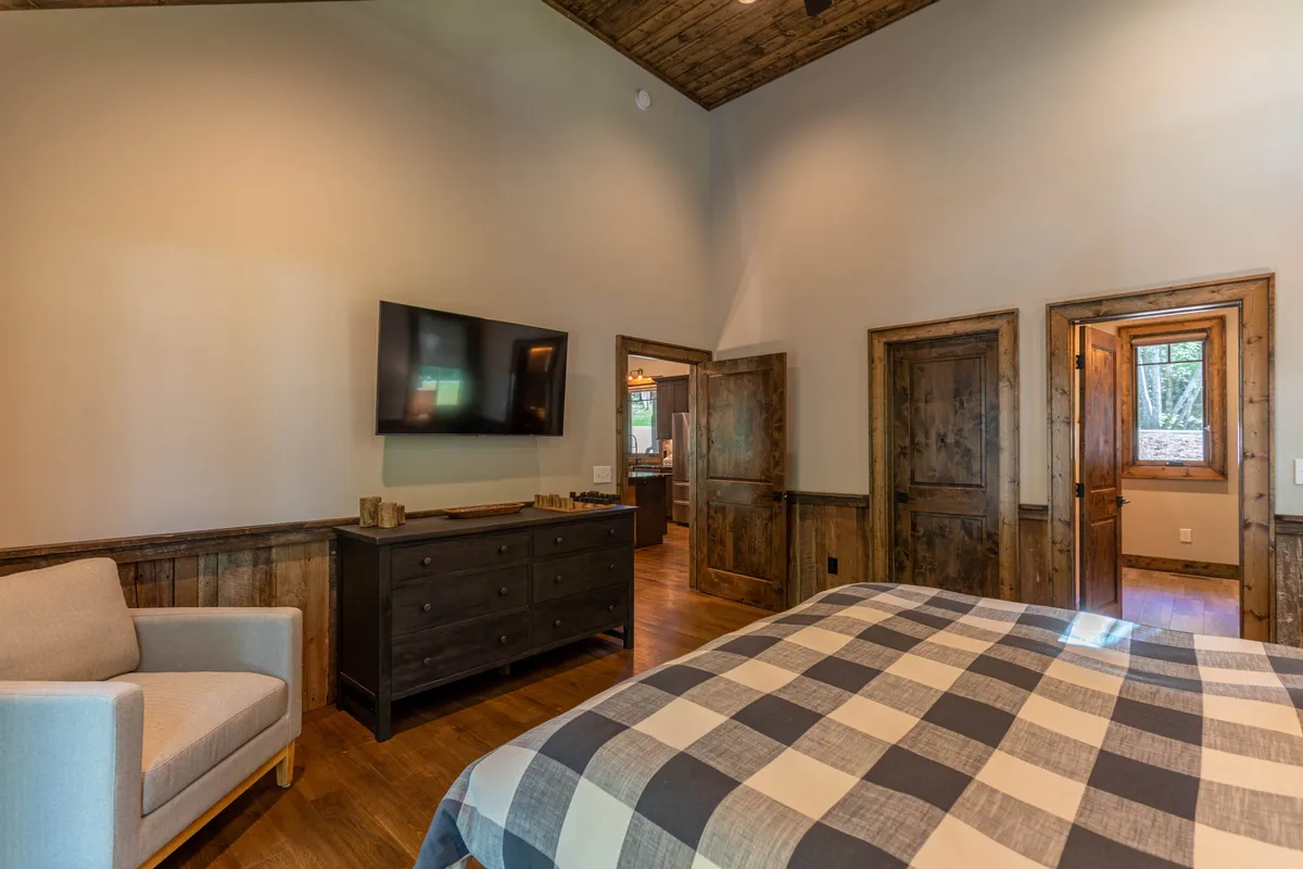 A cozy king bedroom featuring plaid bedding, a wooden dresser, an armchair, and a television. Warm lighting and inviting decor make this suite perfect for unwinding with your favorite shows after a day outdoors.