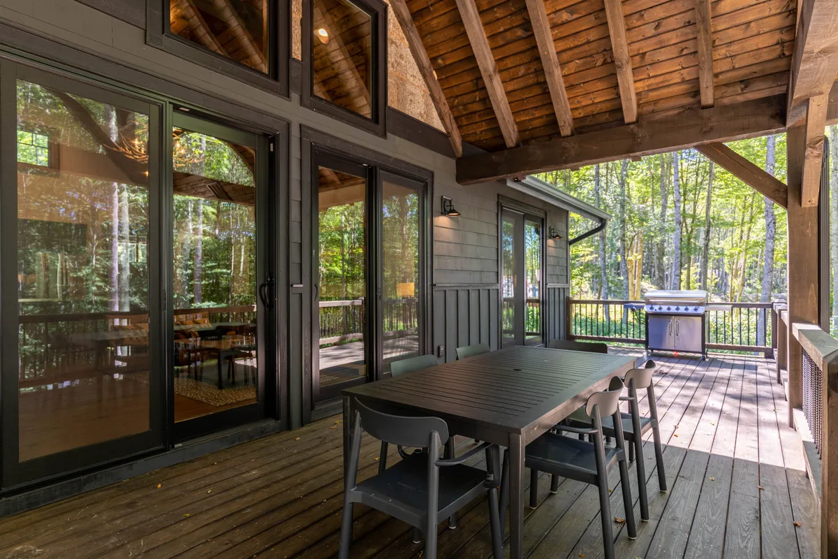 This exterior image features a large dining table set beneath the canopy of surrounding trees, with a grill nearby. The setting exudes a cozy, communal atmosphere perfect for group barbecues and outdoor meals, framed by the natural beauty of the area.