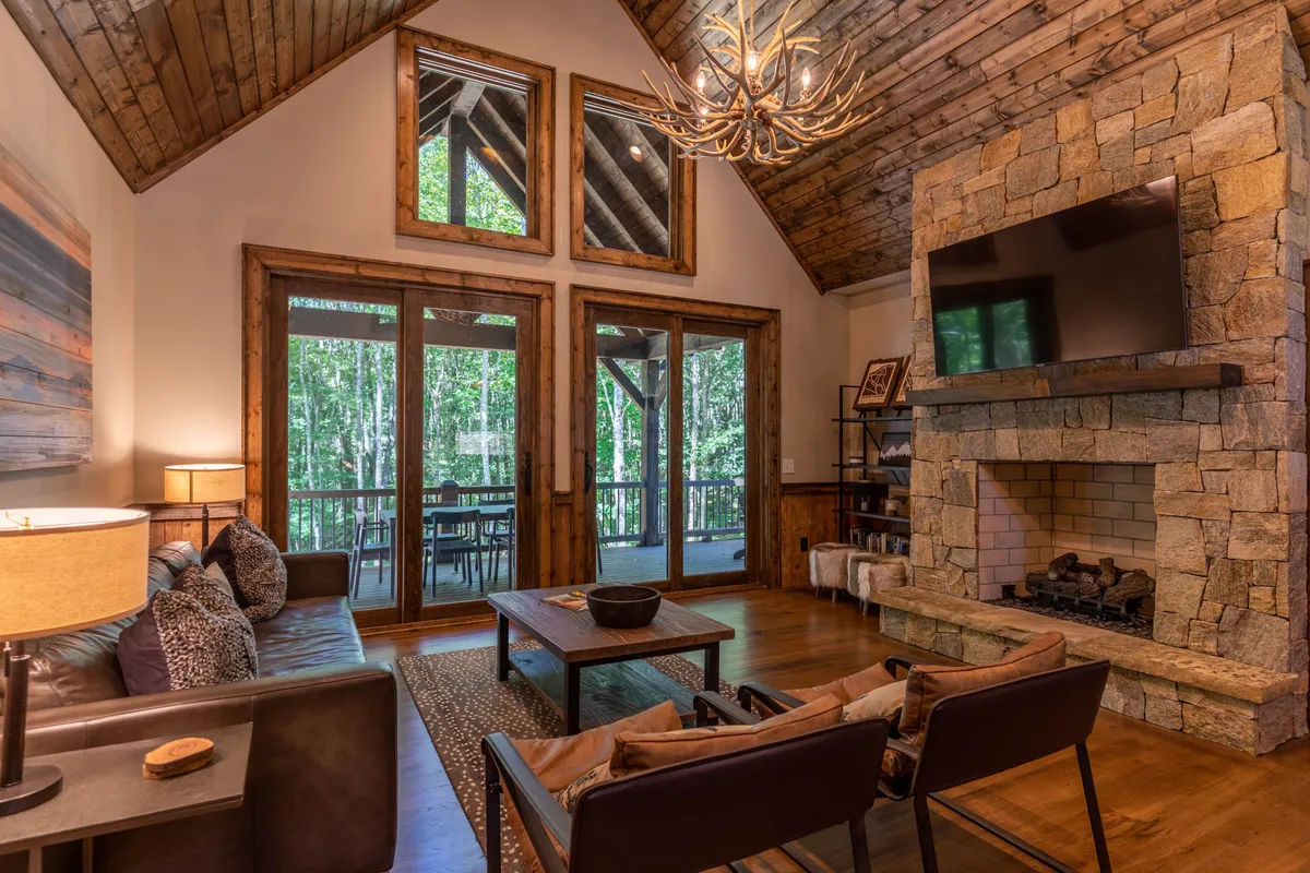 A cozy living space with a plush sofa facing a gas log fireplace, large windows with views to the outdoors, and an elegant chandelier overhead. The room is bathed in natural light, inviting warmth and relaxation for gatherings or quiet evenings.