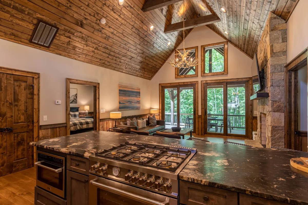 A spacious, sunlit open-concept living area featuring a central kitchen island with a stove, large windows overlooking a scenic natural landscape, striking wooden beams and ceiling, and a prominent stone fireplace. The inviting sofa and dining area are visible, creating a seamless transition between cooking, dining, and relaxing.