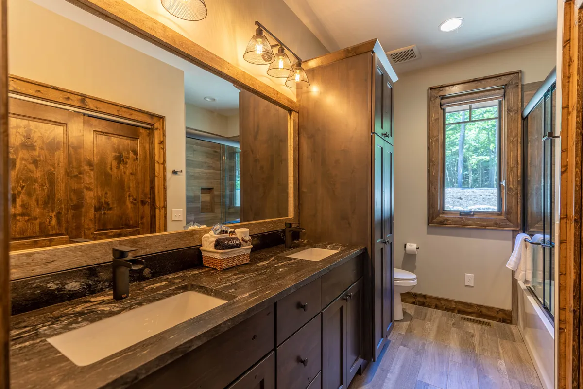 A bright ensuite bathroom with a double sink vanity, large mirror, and natural light pouring in through a window. Wooden cabinetry adds warmth, and the space is designed for both function and relaxation, suggesting a luxurious bath experience.