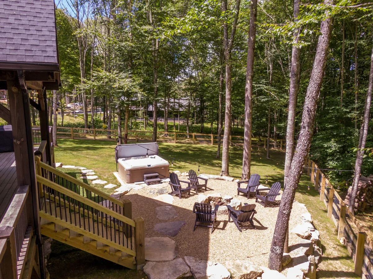 A serene outdoor scene with a hot tub and fire pit as main focal points, complemented by comfortable seating and a backdrop of lush trees. The arrangement encourages both intimate moments and group gatherings, with nature seamlessly blending into the living space.