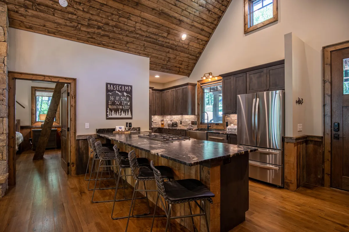 A spacious, warmly lit kitchen with a stone accent wall, a prominent kitchen island, and stainless steel appliances. The wooden ceiling and open layout give a rustic yet upscale cabin feel. While the laundry center and half bath are not directly visible, this image situates viewers in the area adjacent to where these practical amenities are located.