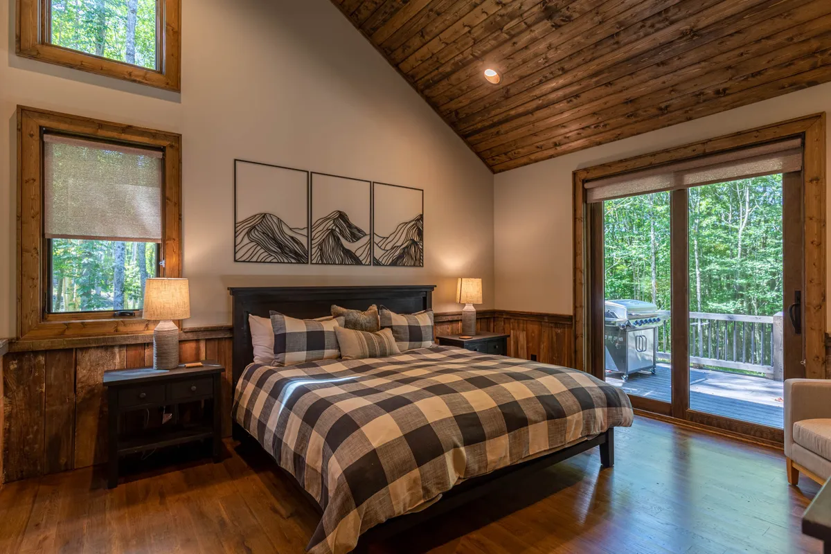 A serene king bedroom with plaid bedding, wooden nightstands, large windows offering a view of the surrounding nature, and mountain art on the wall. The room exudes comfort and openness, with natural light streaming in and a restful atmosphere.