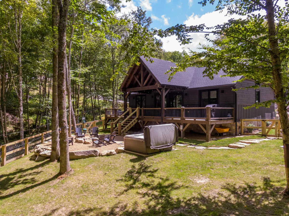 The image showcases a modern mountain cabin nestled in the woods of Banner Elk, North Carolina. The exterior features inviting outdoor seating, a private hot tub, and a spacious deck perfect for stargazing or sipping morning coffee. Surrounded by nature, the home offers a serene retreat ideal for relaxation and entertaining.