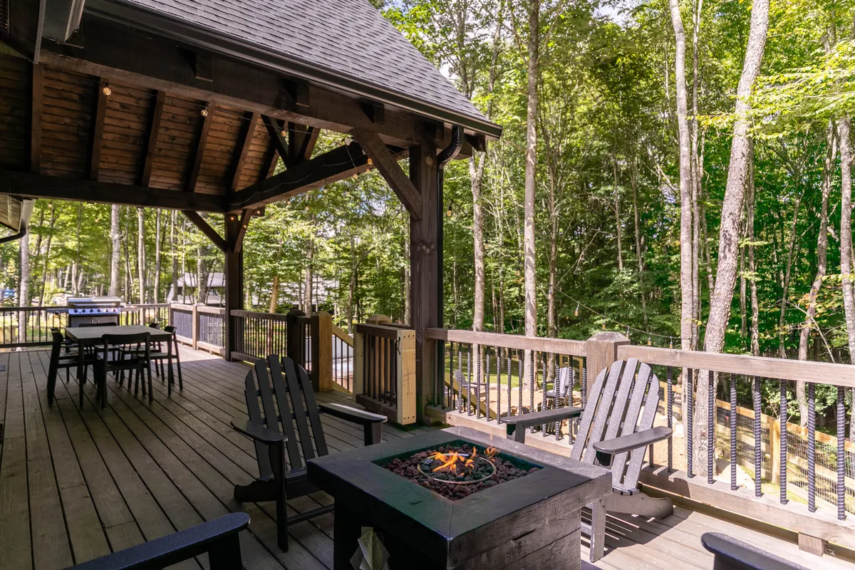 An inviting outdoor seating area arranged around a fire pit on a spacious wooden deck, surrounded by tall trees. The warm glow of sunset filters through the trees, highlighting the intimate arrangement ideal for couples to relax, enjoy a quiet evening, or stargaze by the fire.