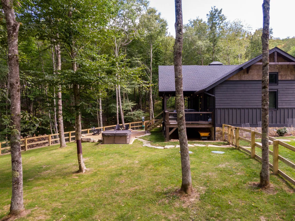 A modern cabin set within a fenced yard, highlighted by a hot tub on the deck and surrounded by greenery. The enclosed lawn space is perfect for pets and children, while the hot tub invites restorative moments amid nature.